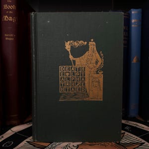 May include: A green book with gold lettering and illustrations. The title of the book is "The Book of the Dead". The cover features a figure holding a torch and a square grid with the word "DEATH" spelled out in gold letters.
