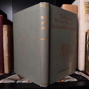 May include: A vintage book titled "The Cave Of The Oracle" with gold lettering on a gray-green cover. The spine displays the title and author, L.W. delaurence. The book is part of a collection, with other books visible in the background.