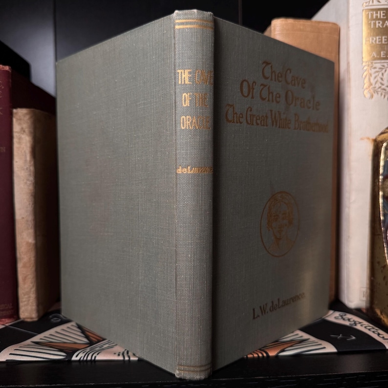 May include: A vintage book titled "The Cave Of The Oracle" with gold lettering on a gray-green cover. The spine displays the title and author, L.W. delaurence. The book is part of a collection, with other books visible in the background.