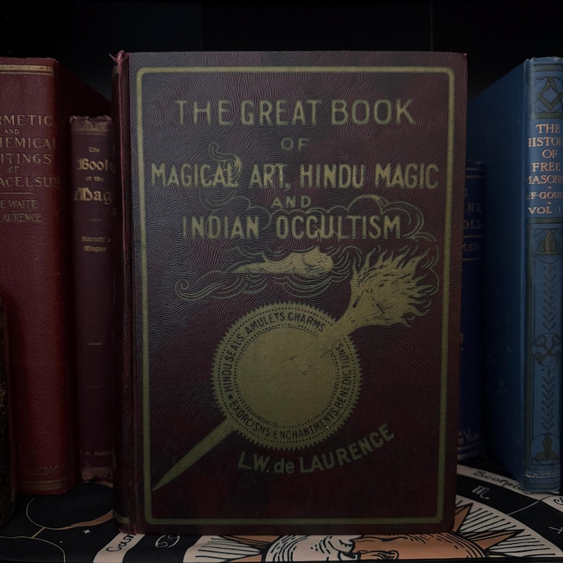 Pu&ograve; includere: Un libro rilegato in bordeaux intitolato "The Great Book of Magical Art, Hindu Magic and Indian Occultism" di L.W. de Laurence. La copertina presenta un'illustrazione dorata di una bacchetta infuocata.
