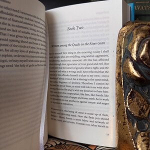 May include: An open book with text on the pages, featuring the title "Book Two" and the beginning of a chapter. A decorative gold-colored book is partially visible on the right side, with the word "AVATS" on the spine of a book in the background.