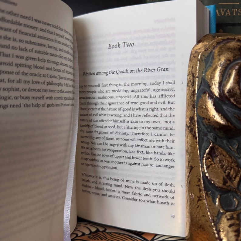 May include: An open book with text on the pages, featuring the title "Book Two" and the beginning of a chapter. A decorative gold-colored book is partially visible on the right side, with the word "AVATS" on the spine of a book in the background.