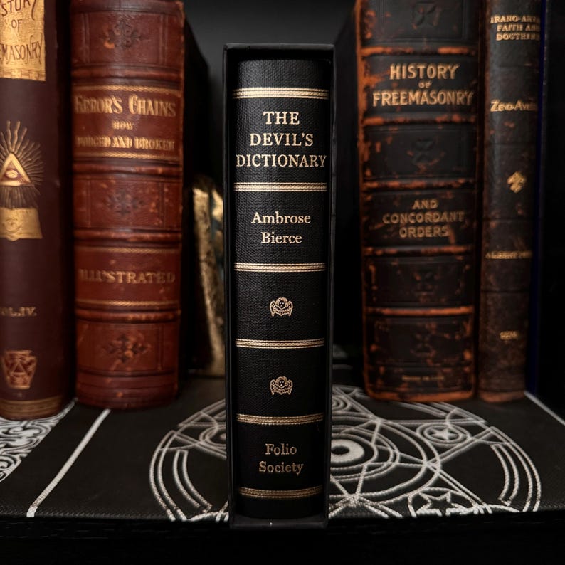 May include: A black leather-bound book titled "The Devil's Dictionary" by Ambrose Bierce. The book is part of the Folio Society collection and features gold lettering and decorative accents.