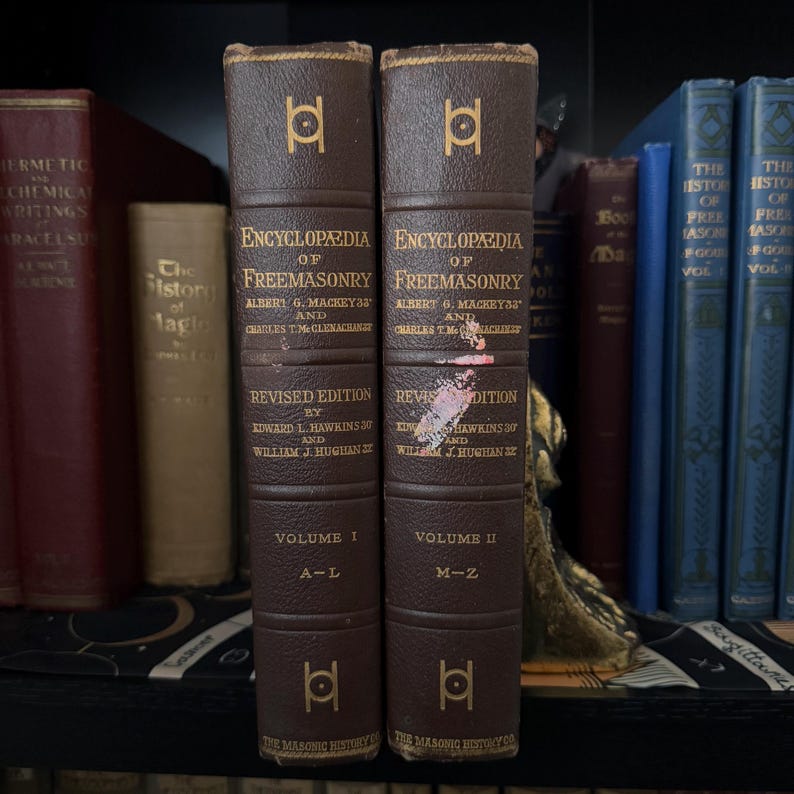 Puede incluir: Dos libros antiguos encuadernados en cuero marr&oacute;n, titulados "Enciclopedia de la Francmasoner&iacute;a", se exhiben en una estanter&iacute;a. Los libros tienen letras y s&iacute;mbolos dorados, con el Volumen I y el Volumen II visibles. Otros libros son visibles en el fondo.
