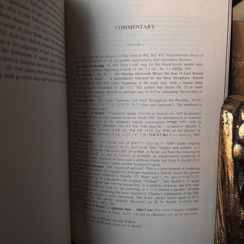 May include: Open book with text titled "COMMENTARY" and "CHAPTER I". The pages are filled with small text, with Greek and Hebrew characters. The book is open on a wooden surface.