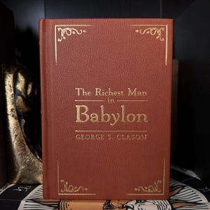 May include: A hardcover book titled "The Richest Man in Babylon" by George S. Clason. The book has a reddish-brown cover with gold lettering and decorative corner designs. The book is displayed on a shelf.