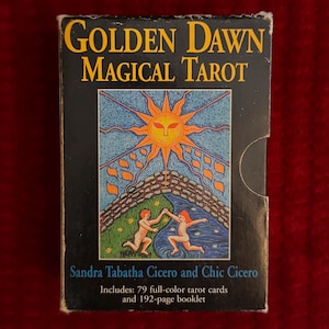 May include: Box for the Golden Dawn Magical Tarot deck. The box is black with gold lettering and features an illustration of a sun, a river, and two figures. The text on the box reads "Golden Dawn Magical Tarot" and "Sandra Tabatha Cicero and Chic Cicero". The box also includes the text "Includes: 79 full-color tarot cards and 192-page booklet".