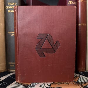 May include: A burgundy hardcover book with a black geometric design on the front cover. The design is a stylized triangle with arrows pointing in different directions.