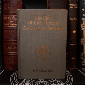 May include: A vintage book titled "The Cave Of The Oracle The Great White Brotherhood" by L.W. de Laurence. The book cover is a light green with a gold embossed design of a woman's head.