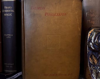 Peut inclure: Un livre vintage intitulé "Demon Possession" avec des lettres rouges sur une couverture beige. Le livre présente un petit motif en relief et le nom de l'auteur en bas. D'autres livres sont visibles sur l'étagère, dont un intitulé "Transcendental Magic".