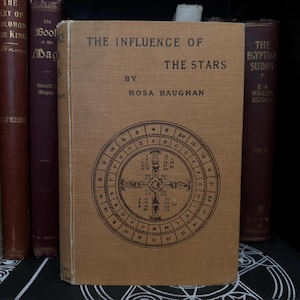 May include: A vintage book titled "The Influence of the Stars" by Rosa Baughan. The cover features a brown background with a black circular design containing letters and numbers.