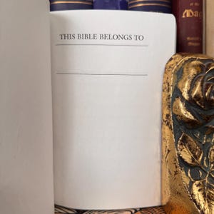 May include: Open Bible with a blank page that reads "THIS BIBLE BELONGS TO" with a line below for the owner's name. The book is surrounded by other books and a decorative gold-colored object.