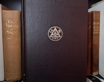 May include: A dark burgundy book with a gold emblem on the cover, featuring a triangle and the word "TRY". The book is standing upright next to other antique books with gold lettering. The book has a textured cover.