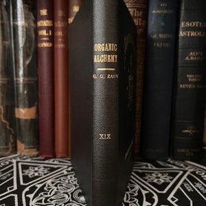 May include: A black leather-bound book titled "Organic Alchemy" by C.C. Zain. The book is part of a collection of books on a shelf. The book has gold lettering and the number XIX on the spine.