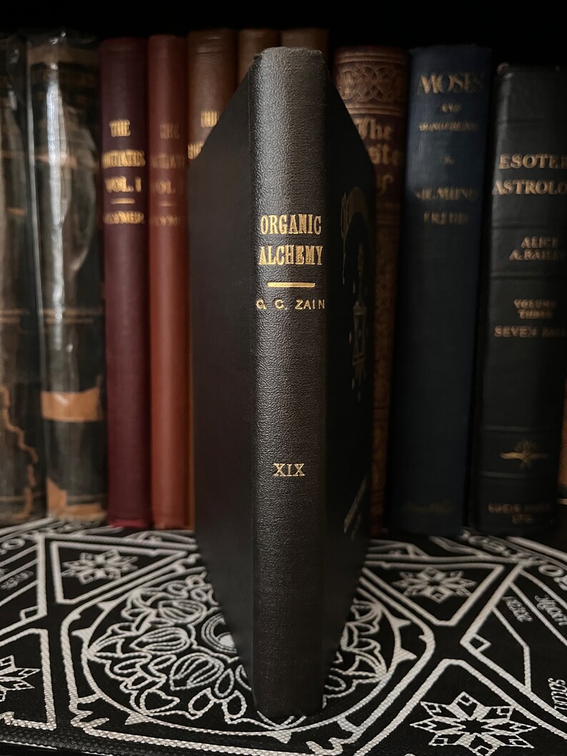 May include: A black leather-bound book titled "Organic Alchemy" by C.C. Zain. The book is part of a collection of books on a shelf. The book has gold lettering and the number XIX on the spine.