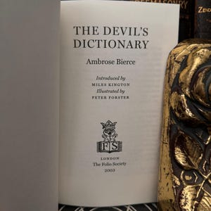 May include: The Devil's Dictionary by Ambrose Bierce, a book published by The Folio Society in 2003. The cover features a black and white illustration of a crowned owl with the initials "FS" in a circle.