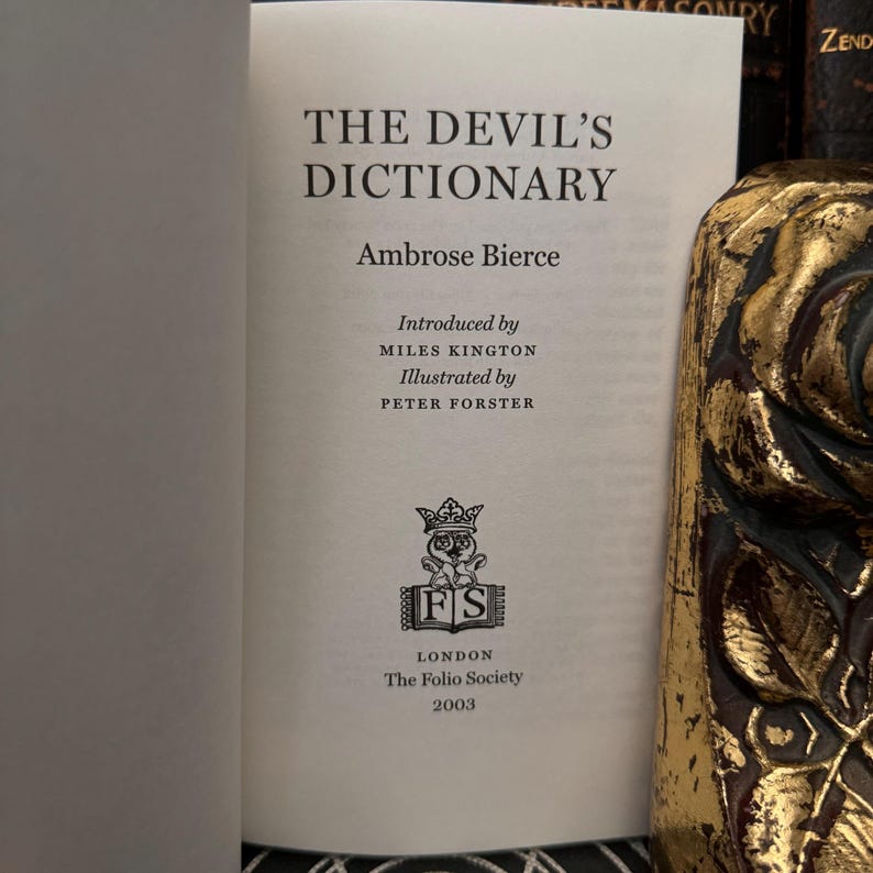 May include: The Devil's Dictionary by Ambrose Bierce, a book published by The Folio Society in 2003. The cover features a black and white illustration of a crowned owl with the initials "FS" in a circle.