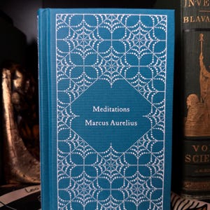 May include: A teal hardcover book titled "Meditations" by Marcus Aurelius. The cover features a white, ornate, dotted design. The book is displayed upright, with the spine visible, showcasing the title and author's name in white text.