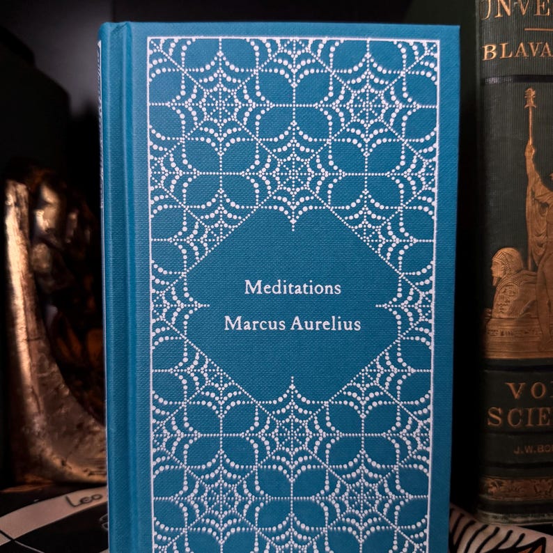 May include: A teal hardcover book titled "Meditations" by Marcus Aurelius. The cover features a white, ornate, dotted design. The book is displayed upright, with the spine visible, showcasing the title and author's name in white text.