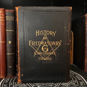 Op de afbeelding: Een zwart lederen gebonden boek met gouden letters met de titel "Geschiedenis van de Vrijmetselarij en de Concordante Orden". De omslag toont een gouden vrijmetselaars symbool.