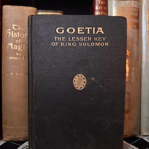 Op de afbeelding: Een zwart boek met gouden letters met de titel "GOETIA THE LESSER KEY OF KING SOLOMON" en een gouden embleem. Het boek maakt deel uit van een verzameling antieke boeken.