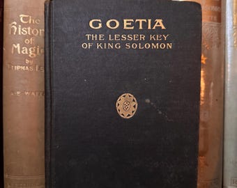 May include: A dark book titled "GOETIA THE LESSER KEY OF KING SOLOMON" with gold lettering and a gold emblem. The book is bound in black cloth and is part of a collection of antique books.