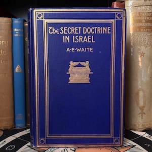 May include: A dark blue hardcover book titled "The Secret Doctrine in Israel" by A.E. Waite, with gold lettering and a gold illustration. Other books are visible in the background, suggesting a collection.