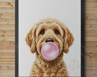 May include: A framed print featuring a golden doodle dog with a pink bubble gum bubble. The dog has brown eyes and a fluffy, light brown coat. The artwork is set against a white background and framed in a black frame, mounted on a wooden wall.