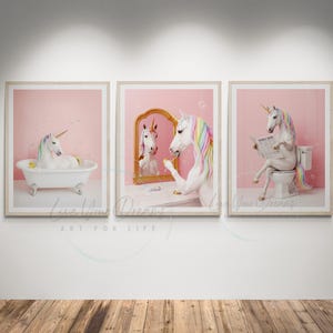 Könnte beinhalten: Ein Set aus drei gerahmten Wandbildern mit einem weißen Einhorn mit regenbogenfarbener Mähne und Schweif. Das erste zeigt das Einhorn in einer weißen Badewanne, das zweite beim Schminken vor einem goldenen Spiegel, das dritte auf einer Toilette.