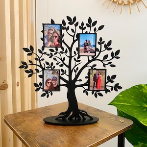 Peut inclure: Cadre photo en métal noir en forme d'arbre avec quatre emplacements pour photos. Le cadre est posé sur une table en bois avec une plante verte en arrière-plan.