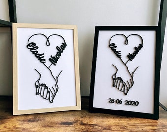 May include: Two framed prints with black silhouettes of hands holding each other, forming a heart shape with the names "Cladie" and "Nicolas" written inside. The date "26 06 2020" is written below the heart on the black frame.