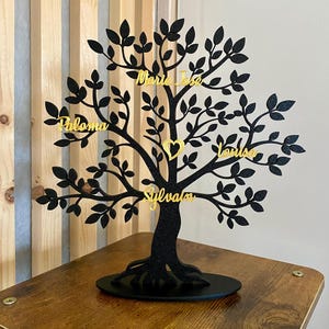 Peut inclure: Silhouette noire d'un arbre avec des accents de paillettes dorées. L'arbre a cinq branches avec des noms écrits dessus : "Paloma", "Maria Jose", "Louise", "Sylvain" et un cœur au centre. L'arbre est posé sur une base noire.