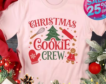 May include: A soft pink t-shirt with a festive Christmas design. The graphic includes the words "CHRISTMAS COOKIE CREW" with images of a whisk, rolling pin, Christmas tree, mitten, and gingerbread person. A "SALE 25% OFF" sticker is visible.