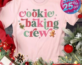 May include: A soft pink t-shirt with the words "Cookie Baking Crew" in a retro font. The design includes a gingerbread man, a spoon with holly, and small cross accents. A "Sale 25% Off" sticker is visible.