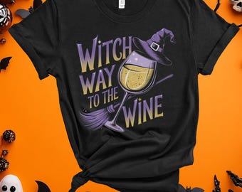 May include: Black t-shirt with a Halloween-themed graphic. The design features a wine glass with a witch's hat, a broom, and the text "Witch Way to the Wine" in purple and yellow. The shirt is set against an orange background.