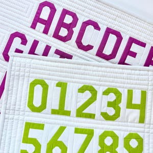 May include: A white quilted fabric panel featuring bright magenta uppercase letters from A to F in the background and lime green numbers from 0 to 9 in the foreground. The fabric has a grid-like stitched texture throughout the entire surface.