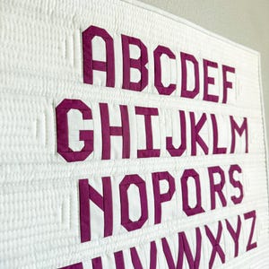Könnte beinhalten: Ein weißer Wandbehang aus Stoff mit dem englischen Alphabet in fetten, magentafarbenen Blockbuchstaben. Die Buchstaben sind auf einen gesteppten weißen Hintergrund appliziert, wobei durchgehend horizontale und vertikale Steppnähte sichtbar sind.