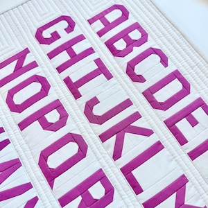 May include: A white quilted fabric panel featuring rows of magenta fabric letters forming the alphabet. The letters are arranged in vertical columns, with the letters A through E, G through L, and N through R clearly visible in a block font style.