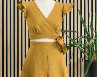 Peut inclure: Une tenue en deux pièces en lin jaune moutarde avec un haut court et une jupe. Le haut a un col en V et des manches à volants. La jupe a un design plissé.