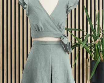 Puede incluir: Un conjunto de top corto y falda de lino verde salvia, con un top estilo cruzado y mangas con volantes. La falda tiene un diseño plisado y un lazo en la cintura.