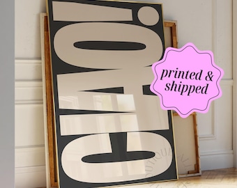 May include: A rectangular wall art print featuring the word CIAO! in large, bold, dark brown sans-serif letters against a light beige background. The print is framed in a thin gold border and rests against a white wall on a wooden floor.