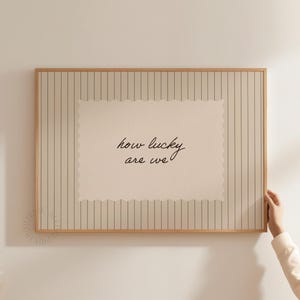 Puede incluir: Impresión enmarcada con el texto escrito a mano "how lucky are we". La impresión tiene un fondo color crema con un borde festoneado y está colocada sobre un fondo a rayas en un marco de madera.