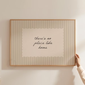 Op de afbeelding: Een ingelijste print met de tekst "there's no place like home" in cursief schrift. Het kunstwerk heeft een crèmekleurige rechthoekige kern met een geschulpte rand, tegen een gestreepte achtergrond in beige en grijs. De lijst is licht hout.