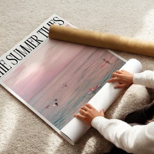 May include: A large poster titled "THE SUMMER TIMES" featuring a pink and blue ocean scene with surfers. A rolled-up poster and a brown cardboard tube are next to the poster. The poster is on a light-colored rug.