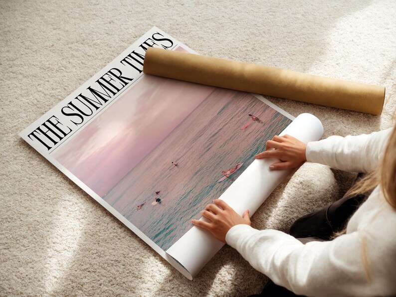 May include: A large poster titled "THE SUMMER TIMES" featuring a pink and blue ocean scene with surfers. A rolled-up poster and a brown cardboard tube are next to the poster. The poster is on a light-colored rug.