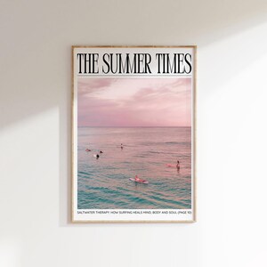 May include: A framed print featuring a serene ocean scene under a pink and purple sky. The text "THE SUMMER TIMES" is at the top. Surfers are in the water. The text "SALTWATER THERAPY: HOW SURFING HEALS MIND, BODY AND SOUL (PAGE 10)" is at the bottom.