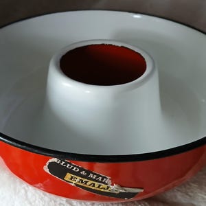 Könnte beinhalten: Eine rot-weiß emaillierte Gugelhupf-Form mit schwarzem Rand. Die Form hat ein zentrales Rohr und ein Etikett an der Seite. Das Innere des Rohrs ist dunkelrot. Ein Vintage-Küchenartikel.