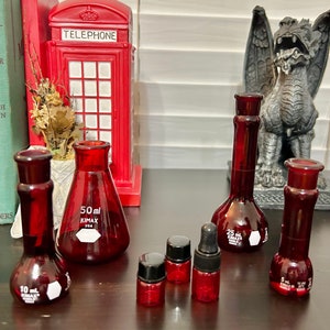 May include: Five red glass bottles with a white label and black text. The bottles are labeled with the text "KIMAX" and the volume in milliliters. The bottles are arranged on a dark brown surface.