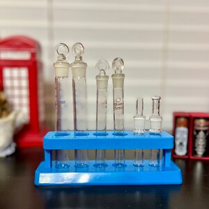 May include: A blue plastic test tube rack with six clear glass test tubes. The test tubes have a white rubber stopper with a small loop on top. The test tubes are labeled with the text "5 ml".