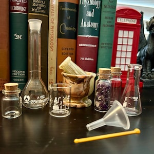 May include: A collection of glass laboratory equipment, including a 25 ml Erlenmeyer flask, a 10 ml Erlenmeyer flask, a funnel, a small glass bottle, and a mortar and pestle. The equipment is arranged on a dark wooden surface with a red telephone booth and a gargoyle statue in the background.